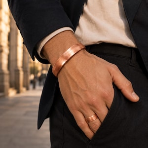 May include: A polished copper cuff bracelet and a matching plain copper band ring. Both pieces feature a smooth, metallic finish with a warm reddish-brown tone, showcasing a simple, unadorned design that wraps around the wrist and the finger.