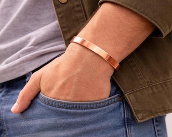 May include: A copper-colored cuff bracelet. The bracelet is a simple, flat band with a polished finish. The image shows the bracelet on a person's wrist, with the person wearing a green jacket and blue jeans.