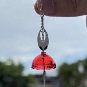 以下が含まれることがあります： チェーン、黒いディテールのある白い楕円形のチャーム、赤いベル型のチャームが付いた、銀色のイヤリング。イヤリングは、ぼやけた緑と空の背景に吊り下げられています。