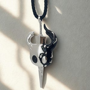 以下が含まれることがあります： 銀と黒のペンダントネックレス。スタイリッシュなキャラクターデザイン。ペンダントは銀と黒の2つに分かれ、顔と尖った刃が描かれています。ネックレスは黒いコードです。