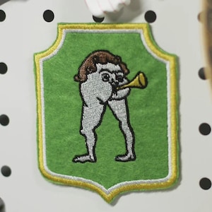 Könnte beinhalten: Gestickter Aufnäher mit einer skurrilen Figur, die Trompete spielt, vor einem grünen Hintergrund mit goldenem Rand. Der Aufnäher ist an einer weißen Lochwand befestigt, zusammen mit dekorativen Vogel- und Blumenmagneten.