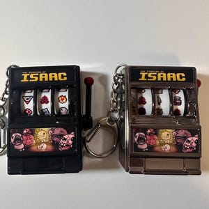 May include: Two slot machine keychains, one black and one silver, with the text "THE BINDING OF ISAAC" across the top. Each keychain has a pull lever and a chain. The slot machine reels display various game-related icons.