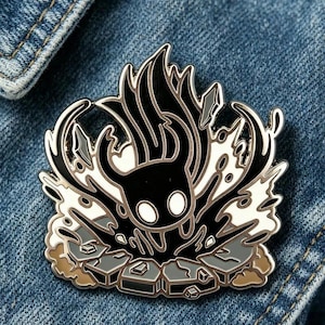 Puede incluir: Un pin de esmalte negro y blanco con un diseño de personaje estilizado y elementos de llama. El pin tiene un contorno plateado y está sobre un fondo de mezclilla. El diseño incluye acentos grises y blancos.