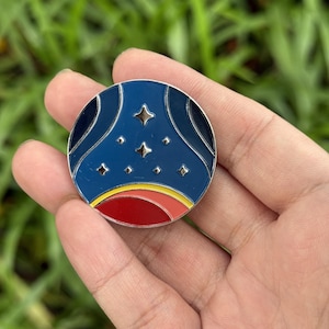 May include: A round enamel pin with a celestial design. The pin features a dark blue background with a stylized planet and stars. The bottom has a red, yellow, and orange band. The pin is held in a hand against a green background.