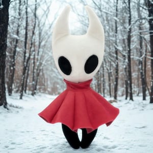 May include: A plush figure of a character with a white head, large black eyes, and two curved horns. The figure wears a red cape and black lower body, standing in a snowy forest setting. The plush toy is a representation of a video game character.