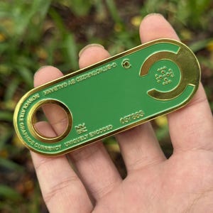 Könnte beinhalten: Ein gold-grüner Metall-Token mit dem Text "NONVOLATILE GRAPHIDE CURRENCY" und "UNIQUELY ENCODED". Der Token hat ein kreisförmiges Loch und ein geschwungenes Design mit kleinen Kreisen. Die Nummer "057883" ist auf dem Token aufgedruckt.