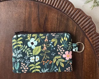 Puede incluir: Un pequeño monedero con cremallera y un estampado floral en tonos verdes, rosas y azules sobre un fondo oscuro. El monedero tiene un llavero y está colocado sobre una bandeja de madera.