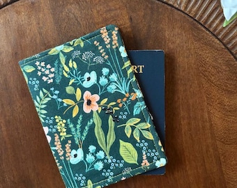 Puede incluir: Un portapasaporte verde oscuro con un estampado floral que presenta flores naranjas, rosas y azules, y hojas verdes. El portapasaporte está sobre una superficie de madera, con un pasaporte azul oscuro parcialmente visible.