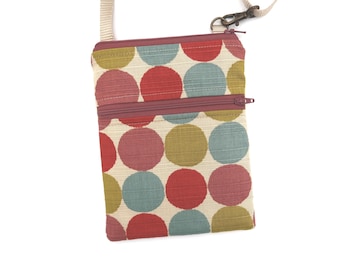 Puede incluir: Un bolso bandolera rectangular de tela color crema con un patrón repetitivo de grandes círculos rojos, verde oliva y azules. El bolso incluye un cierre de cremallera superior, un bolsillo frontal con cremallera y una correa beige ajustable.