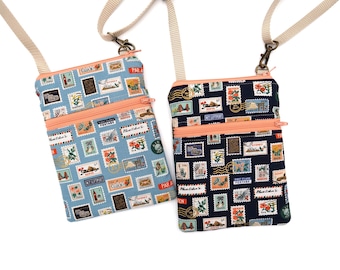 Puede incluir: Dos bolsos bandolera rectangulares con correas ajustables de color beige claro y cremalleras color melocotón. Un bolso tiene un fondo azul claro y el otro un fondo azul marino, ambos con un patrón repetitivo de sellos postales.