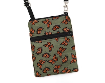 Puede incluir: Un bolso bandolera rectangular con base de tela verde oliva y un patrón repetitivo de mariposas monarca naranjas y negras. El bolso incluye un bolsillo frontal con cremallera negra, ribetes negros y una correa negra ajustable con herrajes metálicos.