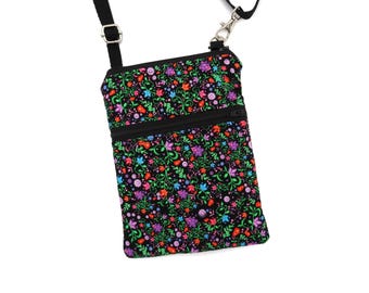 Puede incluir: Un bolso bandolera rectangular con base de tela negra y un estampado floral vibrante en tonos morado, rojo, azul y verde. El bolso incluye un bolsillo frontal con cremallera negra, una cremallera superior negra y una correa negra ajustable.