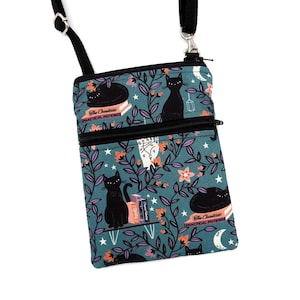 May include: A teal crossbody bag with a black adjustable strap and zipper closure. The fabric features a pattern of black cats, books labeled The Creatures Practical Potions, stars, moons, and leafy vines with orange and purple flowers.