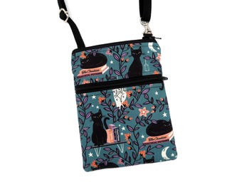 Puede incluir: Un bolso bandolera de color verde azulado con correa negra ajustable y cierre de cremallera. La tela presenta un estampado de gatos negros, libros con el título The Creatures Practical Potions, estrellas, lunas y enredaderas con flores naranjas y moradas.