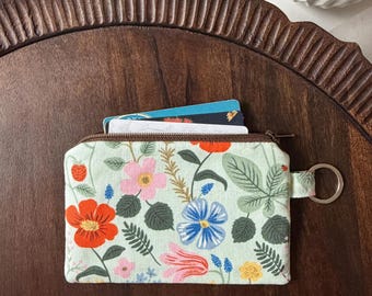 Puede incluir: Un pequeño monedero rectangular con estampado floral, cierre de cremallera y llavero. El monedero presenta un fondo verde claro con un estampado de flores rojas, rosas y azules y hojas verdes. Varias tarjetas son visibles en el interior.