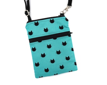 Puede incluir: Un bolso bandolera rectangular de tela color turquesa brillante con un patrón repetitivo de pequeñas siluetas de cabezas de gato negras. El bolso incluye un bolsillo frontal con cremallera negra, un cierre superior de cremallera negra y una correa negra ajustable.
