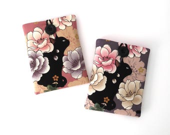 Puede incluir: Dos fundas de tela con estampados florales y siluetas de gatos negros. Cada funda tiene un cierre de botón. Una funda tiene un fondo rosa y la otra un fondo morado. El diseño floral incluye flores rosas, crema y lavanda.