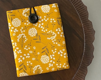 May include: A yellow fabric book cover with a floral pattern of white flowers and dark brown stems. The cover is secured with a black button and elastic loop. The cover is on a dark brown wooden tray.