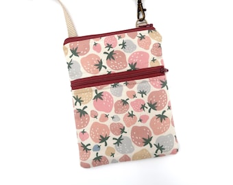 Puede incluir: Un bolso bandolera rectangular con un patrón repetitivo de fresas en tonos rosa, melocotón y azul apagado sobre un fondo crema. El bolso tiene dos compartimentos con cremalleras de color rojo oscuro y una correa beige ajustable.