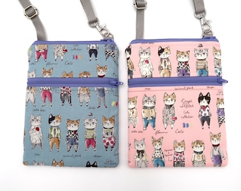 Puede incluir: Dos bolsos bandolera rectangulares con correas grises ajustables y cremalleras moradas. Un bolso es azul claro y el otro rosa claro. Ambos presentan un patrón repetido de gatos ilustrados con diversos atuendos y el texto Rough Sketch Cats collection.