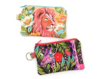 Puede incluir: Dos estuches rectangulares con cremallera y anilla para llaves. El estuche superior muestra un león rodeado de flores coloridas sobre un fondo color melocotón con cremallera turquesa. El inferior muestra una jirafa en un entorno selvático.