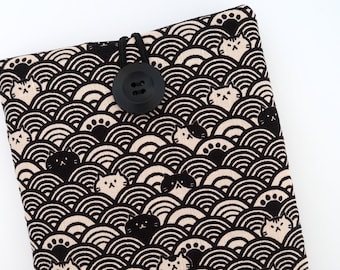 Puede incluir: Una funda de libro de tela en blanco y negro con un patrón de olas estilizadas y gatos. Un botón negro está unido a la funda con un cordón elástico negro.