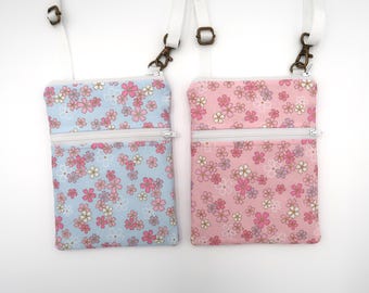 Puede incluir: Dos bolsos bandolera rectangulares con correas ajustables blancas y herrajes de metal en tono bronce. Un bolso presenta una tela azul claro con un patrón floral rosa y blanco, mientras que el otro es rosa con un estampado floral a juego.