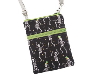 Puede incluir: Un bolso bandolera negro con un patrón repetitivo de esqueletos blancos con sombreros de copa verdes, acentuado con pequeñas estrellas amarillas. El bolso incluye una cremallera de color verde lima y una correa gris ajustable con herrajes de metal plateado.