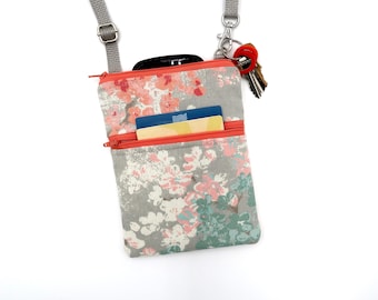 Puede incluir: Un bolso bandolera gris con estampado floral en tonos coral y verde azulado. El bolso cuenta con una cremallera superior, un bolsillo frontal con cremallera que contiene dos tarjetas de crédito y una correa gris ajustable con un clip.
