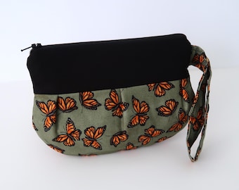 Puede incluir: Una bolsa de cremallera verde y negra con estampado de mariposas. La bolsa tiene una parte superior de tela negra y una parte inferior de tela verde con un patrón de mariposas naranjas. La bolsa tiene un asa de tela negra.