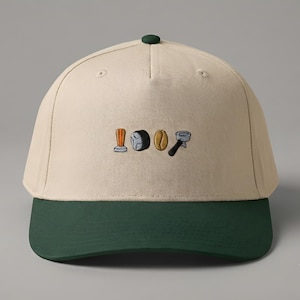 May include: A beige baseball cap with a dark green brim featuring an embroidered design of a coffee tamper, a puck of coffee grounds, a single coffee bean, and a portafilter centered on the front panel of the crown of the hat.