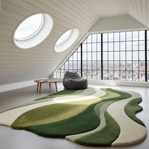 May include: A modern, abstract area rug with a flowing, organic design. The rug features a palette of greens and off-white, creating a layered, wave-like pattern. The rug is placed in a bright, airy room with a beanbag chair and a small wooden table.
