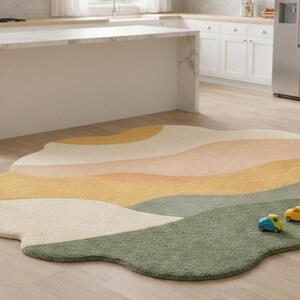 May include: An irregularly shaped area rug featuring a layered, wavy landscape pattern in shades of cream, soft peach, mustard yellow, and sage green. The rug has a plush, textured surface and is accented by two small toy cars in blue and yellow.