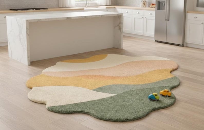 May include: An irregularly shaped area rug featuring a layered, wavy landscape pattern in shades of cream, soft peach, mustard yellow, and sage green. The rug has a plush, textured surface and is accented by two small toy cars in blue and yellow.