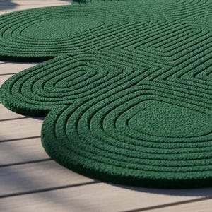 Puede incluir: Una alfombra de color verde oscuro con una superficie texturizada que presenta una serie de formas geom&eacute;tricas redondeadas y conc&eacute;ntricas. La alfombra tiene un contorno irregular distintivo que sigue el patr&oacute;n de las l&iacute;neas en relieve.
