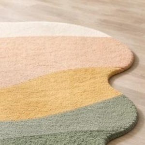 May include: A soft, textured rug with an irregular, organic shape featuring horizontal bands of color in cream, light peach, mustard yellow, and sage green. The surface has a dense, tufted pile construction with a consistent, looped fabric texture.