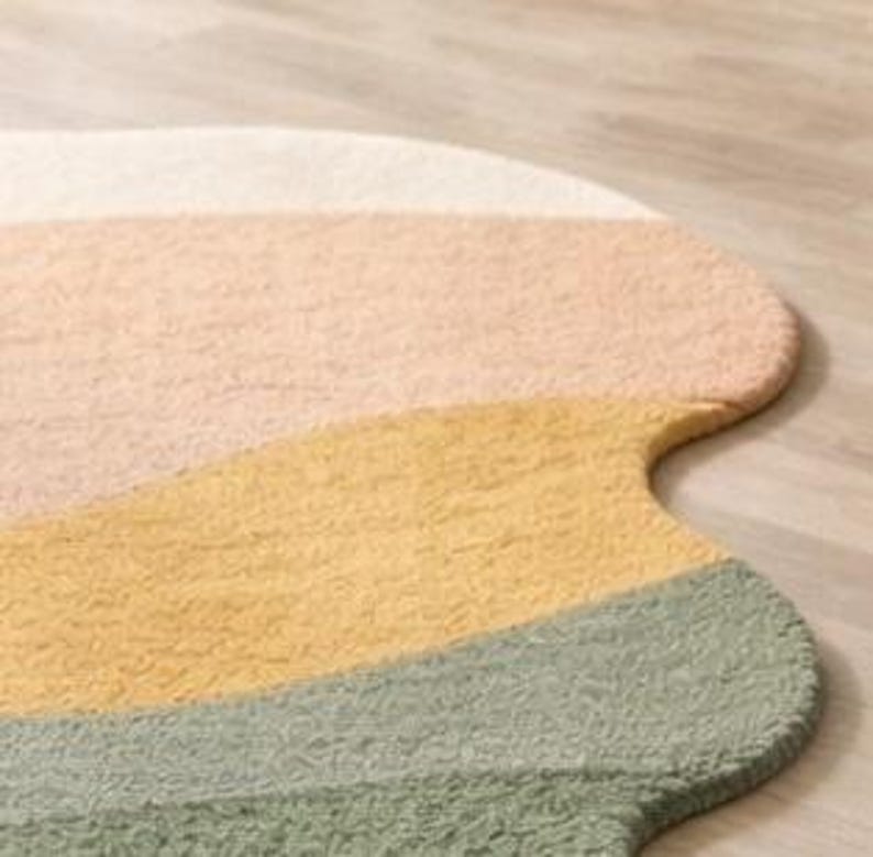 May include: A soft, textured rug with an irregular, organic shape featuring horizontal bands of color in cream, light peach, mustard yellow, and sage green. The surface has a dense, tufted pile construction with a consistent, looped fabric texture.
