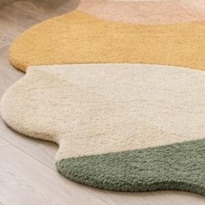 May include: A plush, irregularly shaped area rug featuring a soft, textured pile. The design consists of distinct, curved color-blocked sections in shades of muted peach, warm yellow, cream, and sage green, creating a fluid, organic silhouette.
