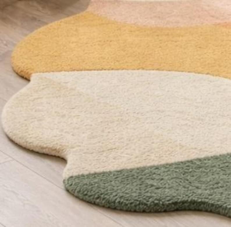 May include: A plush, irregularly shaped area rug featuring a soft, textured pile. The design consists of distinct, curved color-blocked sections in shades of muted peach, warm yellow, cream, and sage green, creating a fluid, organic silhouette.
