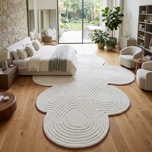 Puede incluir: Una alfombra de color blanco roto con una forma org&aacute;nica grande similar a una nube, que presenta patrones de l&iacute;neas conc&eacute;ntricas talladas. La alfombra est&aacute; colocada sobre un suelo de madera clara, extendi&eacute;ndose bajo una cama por la habitaci&oacute;n.