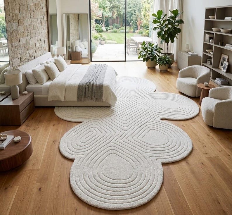 Puede incluir: Una alfombra de color blanco roto con una forma org&aacute;nica grande similar a una nube, que presenta patrones de l&iacute;neas conc&eacute;ntricas talladas. La alfombra est&aacute; colocada sobre un suelo de madera clara, extendi&eacute;ndose bajo una cama por la habitaci&oacute;n.