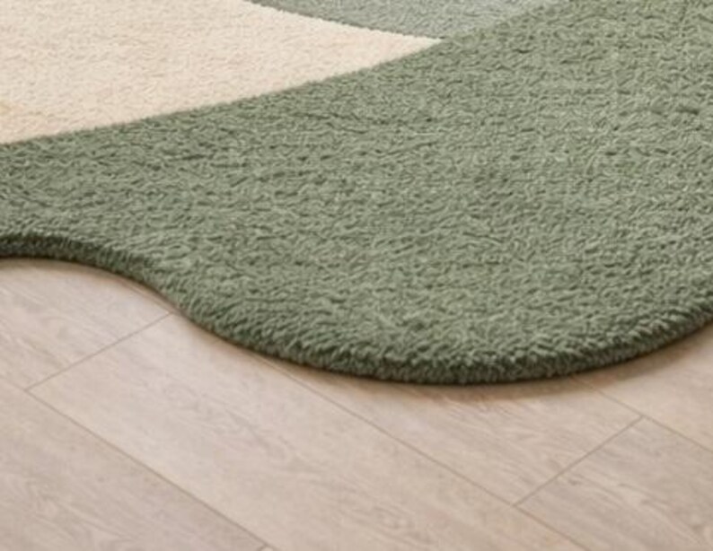 May include: A textured area rug featuring an organic, curved shape with a deep sage green section and a contrasting cream-colored section. The surface has a thick, plush, and curly pile construction that creates a soft, tactile appearance.