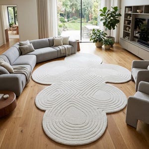 Puede incluir: Una alfombra de color blanco roto con una forma org&aacute;nica &uacute;nica similar a una nube, que presenta patrones de l&iacute;neas conc&eacute;ntricas talladas que siguen los contornos de la alfombra. La alfombra est&aacute; sobre un suelo de madera clara.