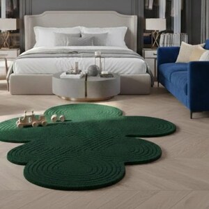 Puede incluir: Una alfombra de color verde oscuro con una forma org&aacute;nica distintiva similar a una nube, que presenta un patr&oacute;n texturizado de c&iacute;rculos conc&eacute;ntricos en toda su superficie. La alfombra se sit&uacute;a sobre un suelo de madera clara en una habitaci&oacute;n.