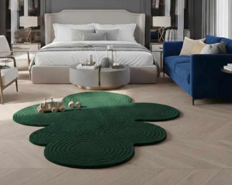 Puede incluir: Una alfombra de color verde oscuro con una forma org&aacute;nica distintiva similar a una nube, que presenta un patr&oacute;n texturizado de c&iacute;rculos conc&eacute;ntricos en toda su superficie. La alfombra se sit&uacute;a sobre un suelo de madera clara en una habitaci&oacute;n.