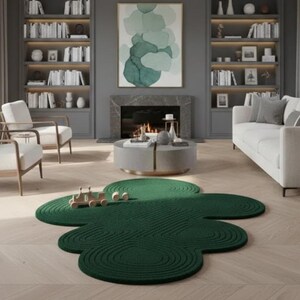 Puede incluir: Una alfombra de color verde oscuro con una forma org&aacute;nica irregular similar a una nube y un patr&oacute;n texturizado de c&iacute;rculos conc&eacute;ntricos. La alfombra est&aacute; colocada sobre un suelo de madera clara en una habitaci&oacute;n con chimenea, estanter&iacute;as y asientos.
