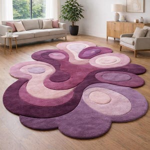May include: A large, abstract area rug in shades of purple, lavender, and cream. The rug features a flowing, organic design with layered shapes and circles. The rug is made of a soft, plush material and is placed on a hardwood floor.