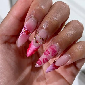 May include: Long, pointed acrylic nails with a pink and red gradient design. Some nails feature heart and pearl accents, while others display a kiss print. The nails have a glossy finish.