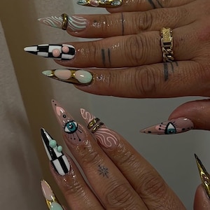 Peut inclure: Gros plan sur de longs ongles pointus avec divers motifs. L'un des ongles pr&eacute;sente un motif en damier noir et blanc, tandis que d'autres affichent des volutes, des motifs d'yeux et des accents dor&eacute;s. Les ongles sont peints dans des tons de rose, d'or et de turquoise.