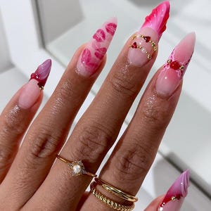 May include: A hand displaying long, almond-shaped acrylic nails. The nails are decorated with designs such as pink lips, red hearts, and gold detailing. Two gold rings are visible on the ring finger.